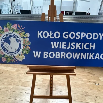 Tablica reklamowa Koło Gospodyń Wiejskich w Bobrownikach z niebieskim tłem i herbem