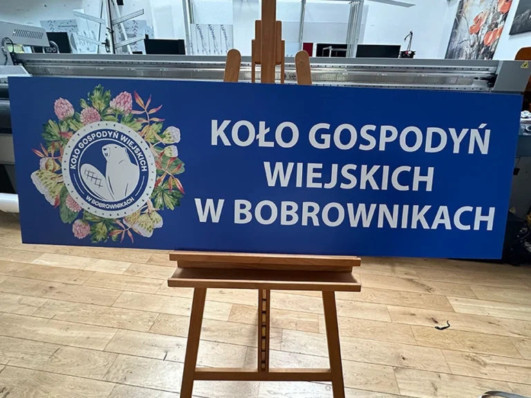 Tablica reklamowa Koło Gospodyń Wiejskich w Bobrownikach z niebieskim tłem i herbem