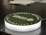 Okrągły kaseton LED z plexi z zielonym frontem i białą typografią, ujęcie boczne, druk UV