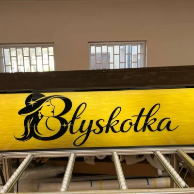 Podświetlany kaseton „Błyskotka” z logo kobiety w kapeluszu i czarnym napisem na żółtym tle