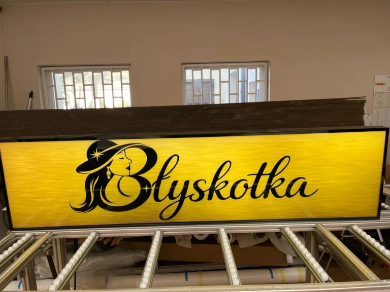 Podświetlany kaseton „Błyskotka” z logo kobiety w kapeluszu i czarnym napisem na żółtym tle