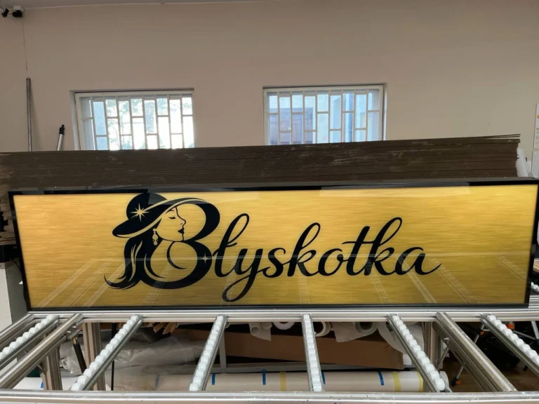 Szyld LED „Błyskotka” na żółtym tle, czarna typografia i logo, ujęcie z przodu w pracowni