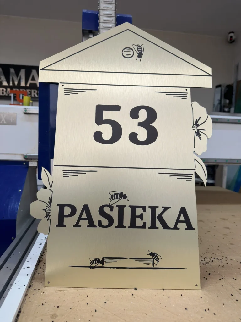 Złoty szyld adresowy w kształcie ula z numerem 53, napisem Pasieka i motywem pszczół