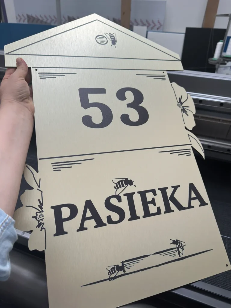 Szyld dla pasieki z numerem 53, czarnym nadrukiem i wycinanym kształtem domu na złotej płycie