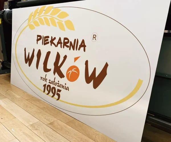 Reklamowy szyld z motywem zboża i nazwą piekarni, wykonany na białej płycie kompozytowej